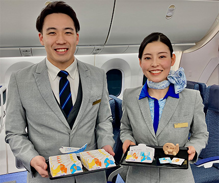 画像：「ANAオリジナルデザイン 明治 ザ・カカオ ナッティカカオ」「同 フルーティ カカオ・ラテ」を持つ客室乗務員